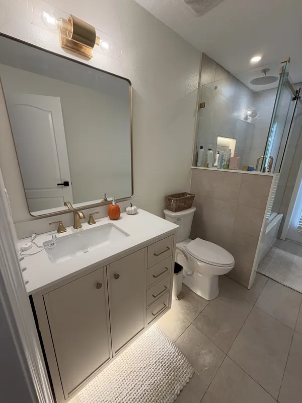 Bathroom with a white vanity, brushed-gold faucet, large rectangular mirror, toilet and glass shower, with a clean tile floor.