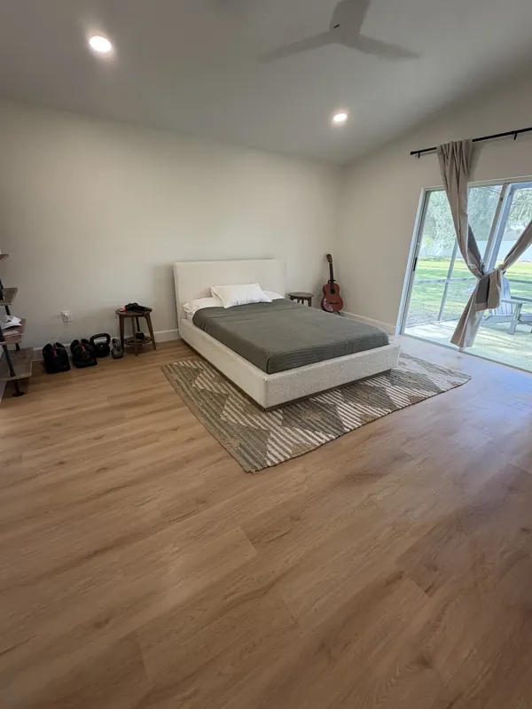 Spacious bedroom with a made bed, area rug and light wood-look floor, sunlight coming through sliding glass doors.
