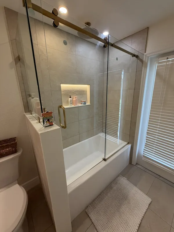 Bathroom with a glass-door shower over a white tub, lit wall niche and gold fixtures, with a white bath mat on the tile floor.