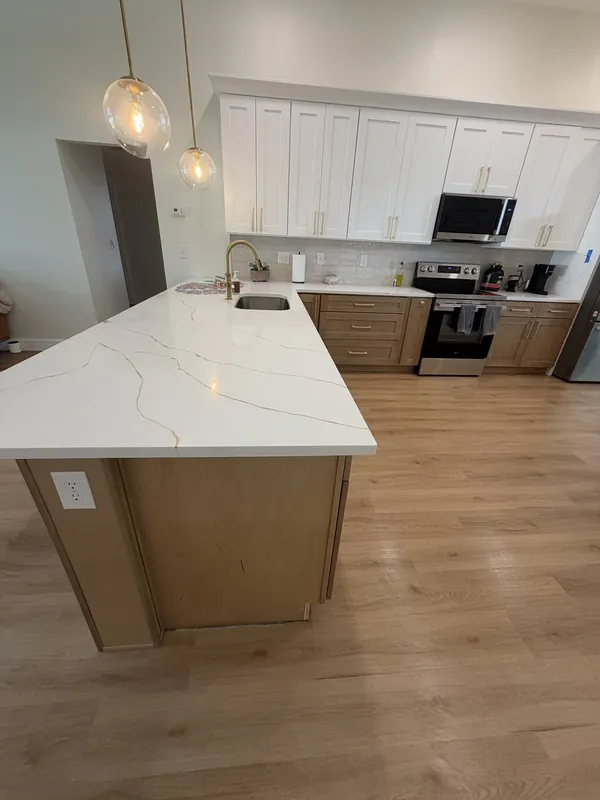Kitchen with a long white-veined quartz island, white upper cabinets, light wood lower cabinets, pendant lights and light wood-look floor, all surfaces clear and clean.