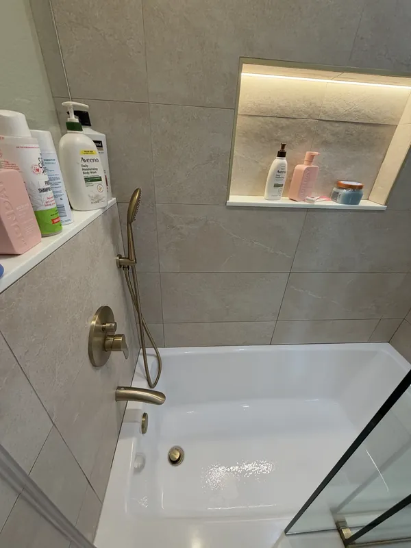 Tub and shower with a lit wall niche holding toiletries, gold handheld shower and clean white tub.