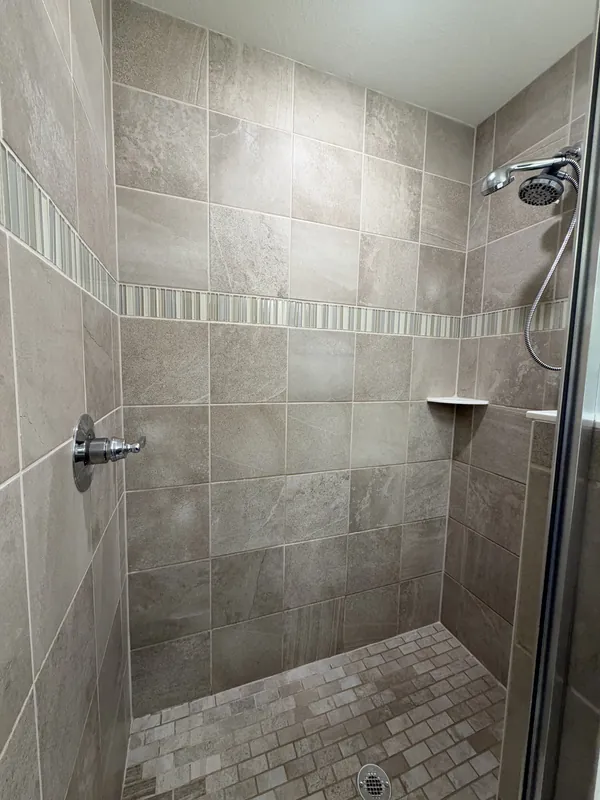Walk-in shower with gray porcelain wall tile, a mosaic accent strip, a corner shelf and a mosaic tile floor, clean and empty.