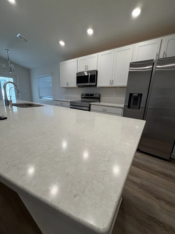 White shaker-cabinet kitchen with a polished light quartz island, stainless refrigerator and microwave, and recessed lights reflecting on the countertop.